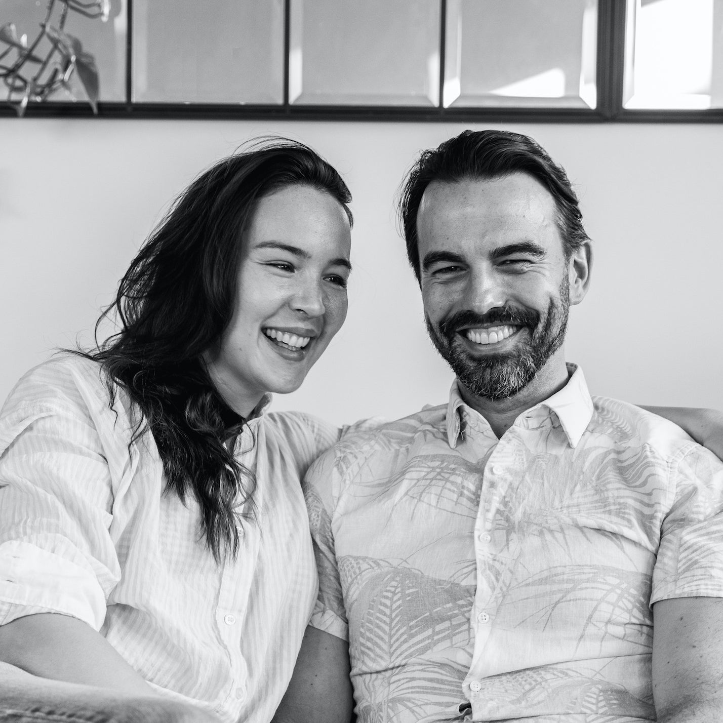 Black and white image of Amanda Pakes and Jordan Pakes sitting on a couch. Image if from the waist up, with both Amanda and Jordan smiling. 