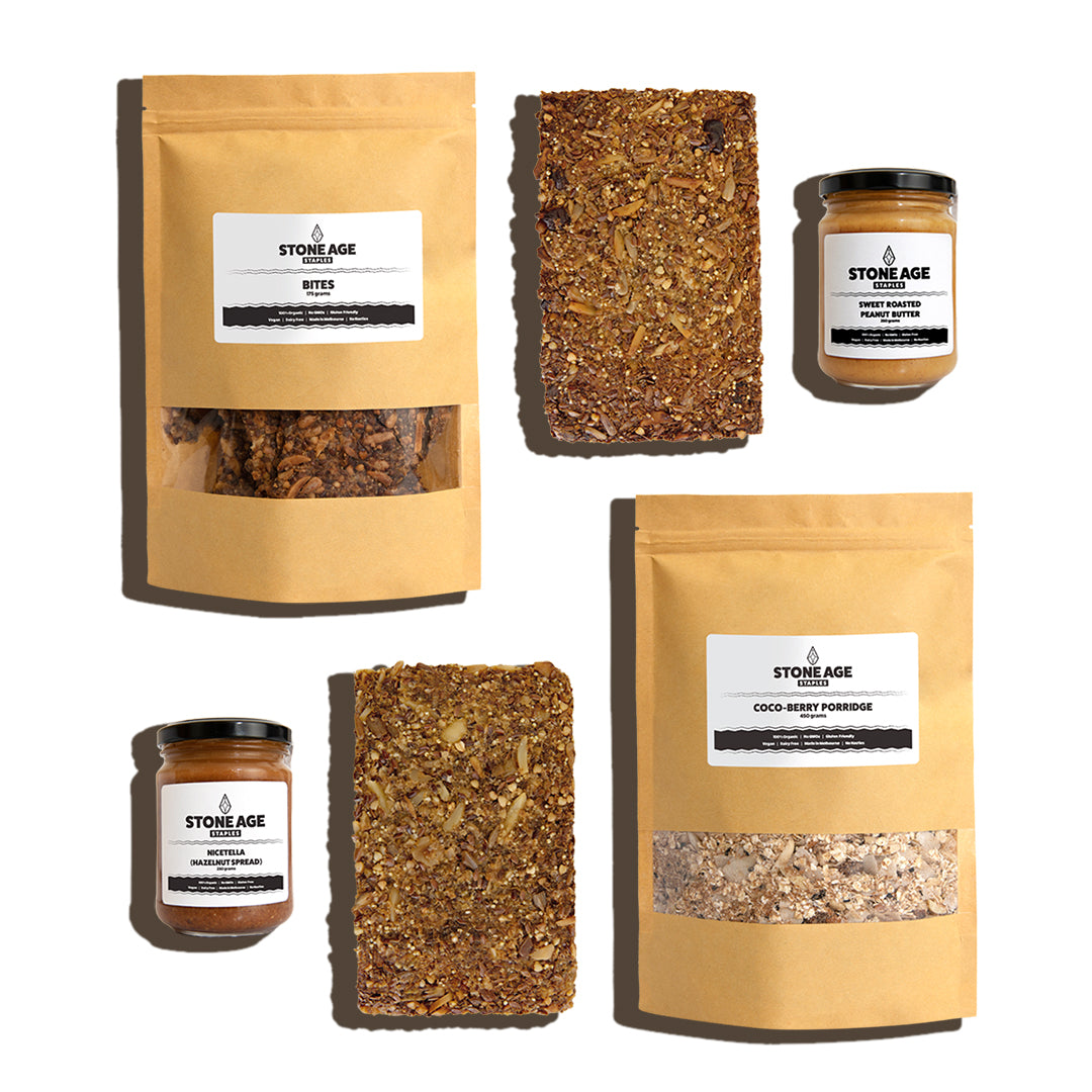 Staple Box. Flat lay image of six products. Two nut grain and seed loaves (one fruit), two nut butters, one packet of bites and one packet of porridge. Displayed on a white background.