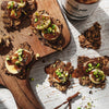 Six bites topped with healthy vegan hazelnut spread, sliced banana and pistachios next to a jar of Nicetella on a wooden chopping board white timber background