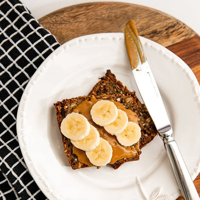 Flat lay of two slices of nut, seed and grain loaf with sweet roasted peanut butter and sliced banana on top. Sitting on a white plate with a silver knife, with the plate sitting on a chopping board with a napkin and white background.