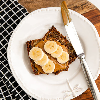 Two slices of Fruit Loaf covered in sweet roasted peanut butter with six slices of banana next to a knife smeared in peanut butter presented on a white plate with a black and white grid tea towel and a wooden board in the background