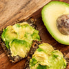 Two slices of Stone Age Staples Original Loaf topped with avocado next to  half an avocado on a wood background