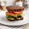 Two slices of Original Loaf presented like sandwich with sliced tomato, tofu, lettuce and avocado inside the sandwich presented on a white textured plate on a grey and white grid tea towel